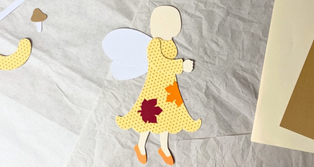 Waldfee aus Papier mit angeklebtem Vorderarm und bunten Ahornblättern auf dem gelben Kleid
